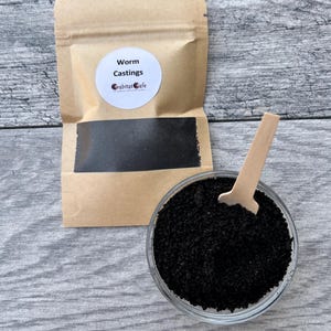 May include: A clear glass bowl filled with dark, granular worm castings, with a small wooden scoop. A brown paper bag with a white label that reads "Worm Castings" and "habitat cafe" is in the background.