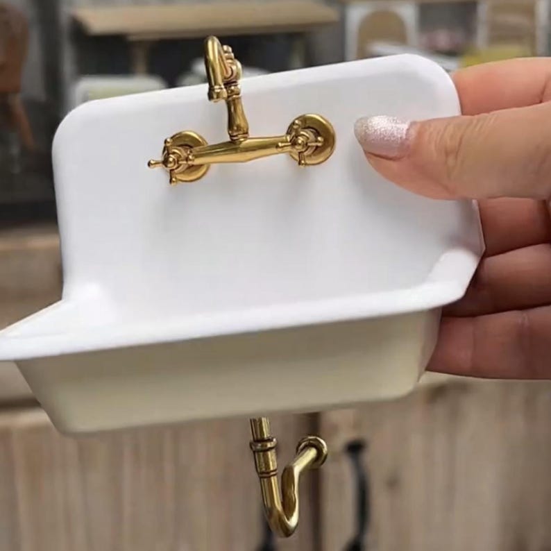 May include: A miniature white porcelain sink with a gold-coloured tap and drainpipe. The tap has a classic design with two handles and a curved spout. The sink is held by a person.