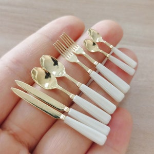 May include: A set of miniature gold-colored cutlery with white handles, including a fork, two spoons, and two knives.