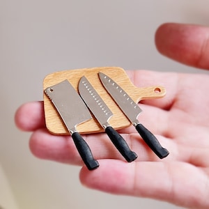 May include: A set of three miniature kitchen knives on a wooden chopping board. The knives have black handles and silver blades. The chopping board is brown with a light wood grain.