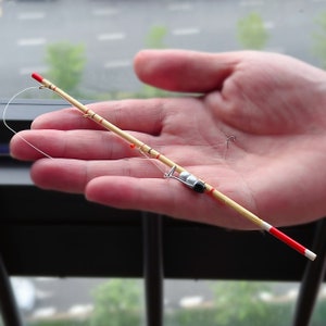 May include: A miniature fishing rod made of bamboo, with red and white detailing. The rod is held in a hand, highlighting its small size. The fishing line and hook are also visible. The rod is approximately 15 cm long.