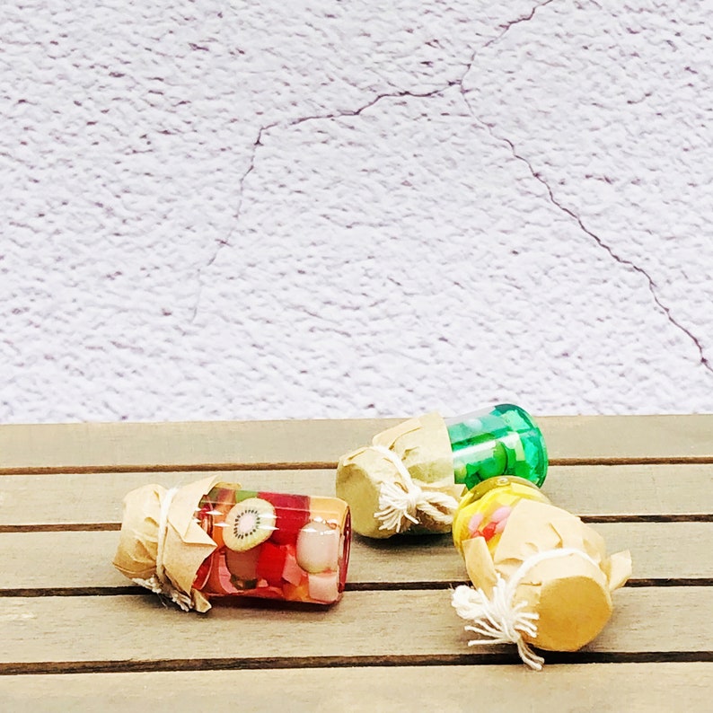 Pu&ograve; includere: Tre piccoli barattoli di vetro con coperchi avvolti in carta marrone e spago. I barattoli sono pieni di caramelle e frutta colorate. I barattoli sono su una superficie di legno.
