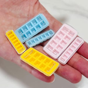 May include: A selection of colourful ice cube trays in various sizes. The trays are yellow, blue, and pink, with clear plastic lids. The trays are designed to make square ice cubes.