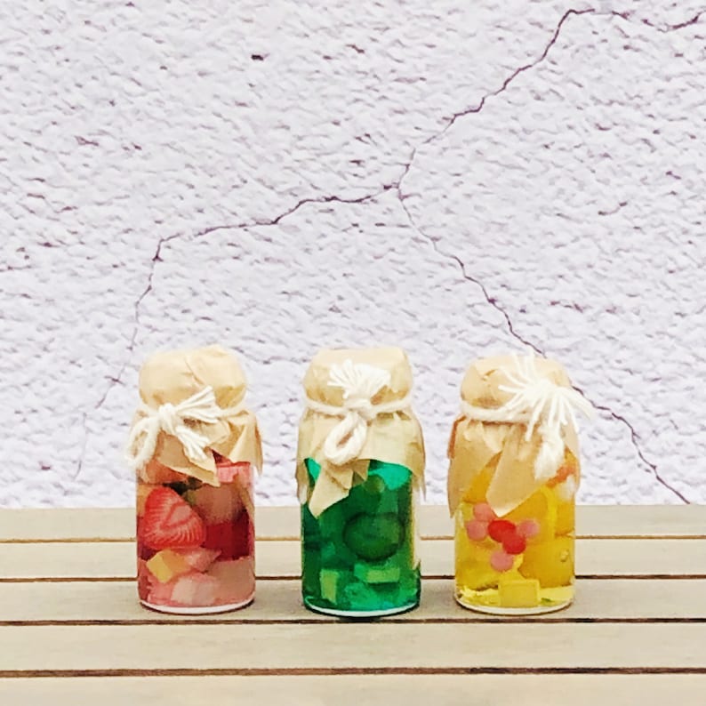 Pu&ograve; includere: Tre piccoli vasetti di vetro con coperchi e rivestimenti in carta marrone legati con spago. I vasetti contengono confetture di frutta colorate. I vasetti sono disposti in fila su una superficie di legno.