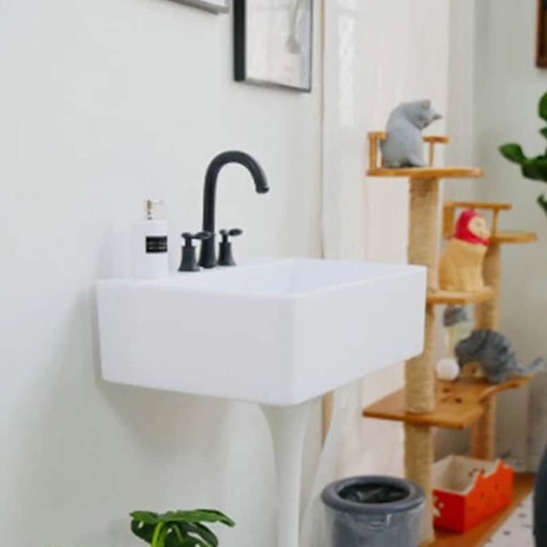 May include: A white pedestal sink with a black faucet and two handles. The sink is in a bathroom with a white wall and a wooden shelf with a cat tree.