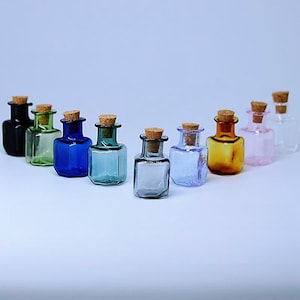 Op de afbeelding: Tien kleine vierkante glazen flesjes met kurkstoppen. De flesjes zijn in verschillende kleuren, waaronder zwart, groen, blauw, teal, grijs, lichtblauw, amber en roze.