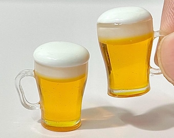 May include: Two miniature beer mugs filled with golden liquid and topped with white foam. The mugs have clear handles and are likely for a dollhouse or miniature collection.