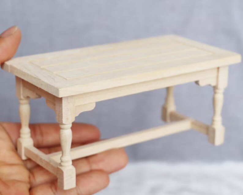 May include: A miniature wooden table with a simple design. The table has a rectangular top and four legs with turned details. The table is made of unfinished wood and is ready to be painted or stained.