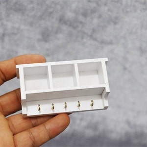 May include: White wooden shelf with three compartments and five hooks. The shelf is designed for a dollhouse or miniature setting.