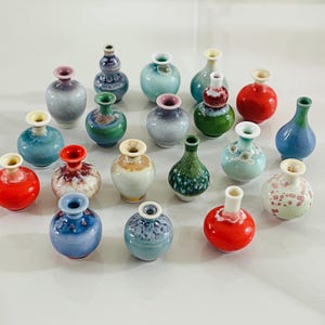 May include: A collection of small, decorative ceramic vases in various colors, including red, blue, green, and white. Each vase has a unique shape and design, with a glossy finish. The vases are arranged on a white surface.