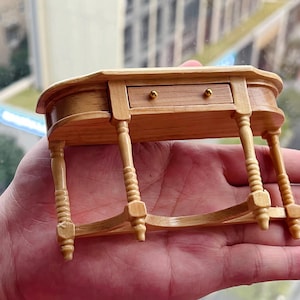 May include: A miniature wooden table with a single drawer and turned legs. The table is painted in a light wood colour and has gold-coloured drawer pulls.
