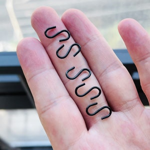 May include: A hand holding a set of six black metal S-shaped hooks.