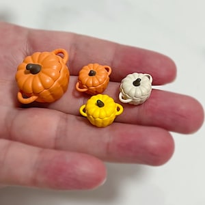 以下が含まれることがあります： 異なるサイズのかぼちゃの形をしたミニチュアのセラミックポットが4つあります。一番大きなポットはオレンジ色、次の2つは黄色とオレンジ色、一番小さなポットは白です。