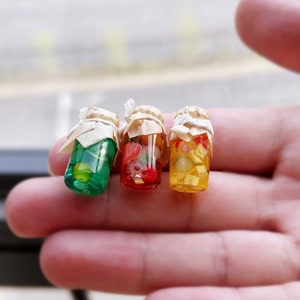 Op de afbeelding: Drie miniatuur glazen potten met deksels, gevuld met kleurrijk fruit. De potten zijn vastgebonden met touw en hebben een rustieke uitstraling.