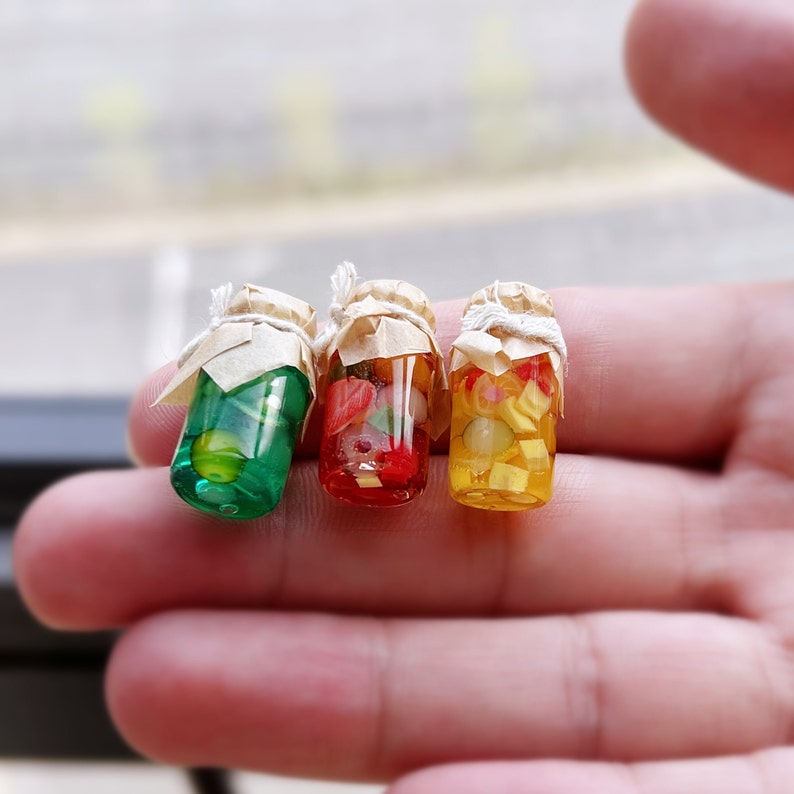 Pu&ograve; includere: Tre vasetti di vetro in miniatura con coperchi, pieni di frutta colorata. I vasetti sono legati con lo spago e hanno un aspetto rustico.