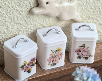 May include: Three white square canisters with silver handles and floral designs. One features pink roses, another has yellow and pink flowers, and the third has purple flowers. A white ceramic bunny is mounted on the wall above.