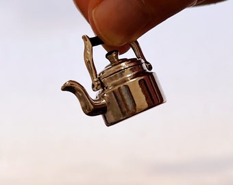 May include: A miniature silver-colored teapot with a curved spout, handle, and lid. The small kettle is held up against a light background. The teapot is a decorative item.