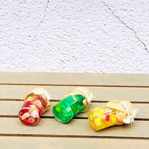 Pu&ograve; includere: Tre vasetti di vetro in miniatura con tappi di sughero e contenuto colorato. I vasetti sono pieni di caramelle rosse, verdi e gialle. I vasetti sono su una superficie di legno.