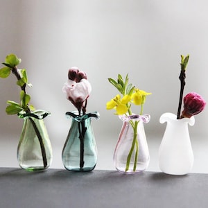 May include: Four small glass vases with different coloured glass and flowers in them. The vases are on a grey surface.