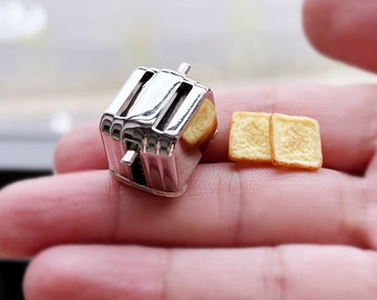 Könnte beinhalten: Ein Miniatur-Toaster aus Silber mit zwei Miniatur-Toastscheiben. Der Toaster und das Toastbrot liegen auf der Hand einer Person.