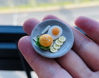 May include: A miniature gray plate with a miniature hamburger, fried egg, and lemon slices. The plate is held in a person's hand.