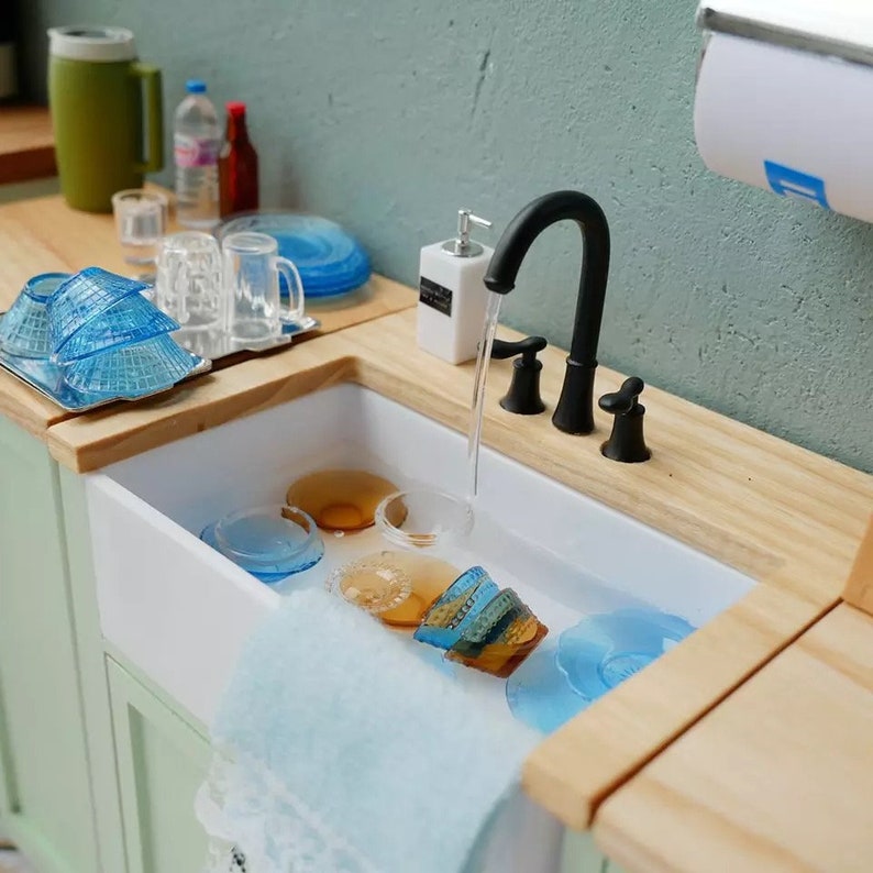 May include: A white porcelain kitchen sink with a black faucet and a blue dish towel hanging over the edge. The sink is filled with water and several blue and brown glass dishes are soaking in the water.