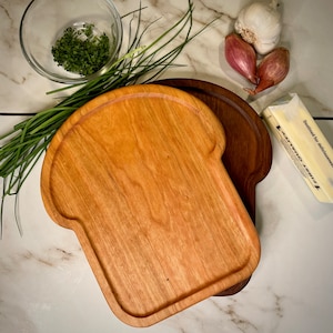 May include: Two wooden serving boards, one shaped like a slice of bread, the other a rounded rectangle. The bread-shaped board is made of light wood, the other is dark wood. Both boards are on a white marble surface.