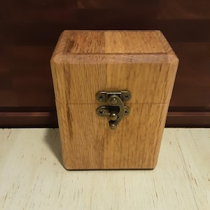 May include: A rectangular wooden box with a natural light brown wood grain finish. The box features a hinged lid secured by a central antique brass-colored metal latch mechanism with visible screws, providing a functional closure for the container.