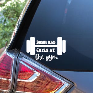May include: White vinyl decal with the text "Down Bad Cryin at the gym" in a stylized font, with a barbell graphic. The decal is affixed to a car window, with the taillight visible in the lower left corner.