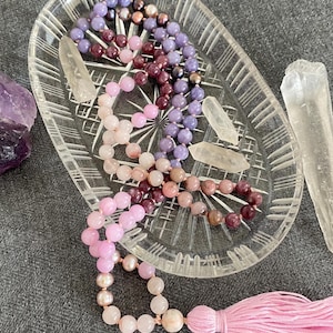May include: A beaded necklace with a pink tassel rests on a clear, etched glass dish. The necklace features a gradient of pink, purple, and brown beads. Clear quartz crystals and a purple amethyst crystal are also present.