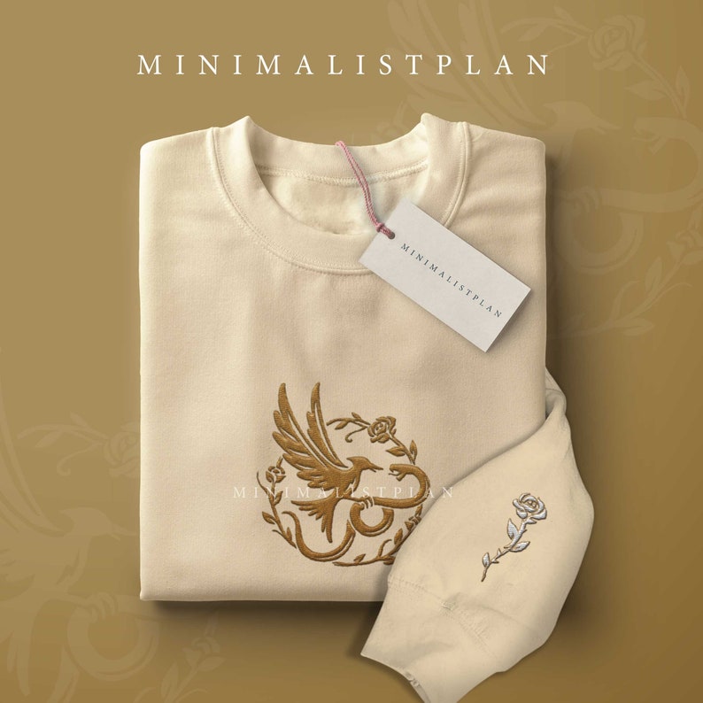 A beige sweater with an imprint of the Hunger Games: Ballad of Songbirds and Snakes symbol on the front and a rose imprint on the sleeve. The sweater sits against a dark tan background.