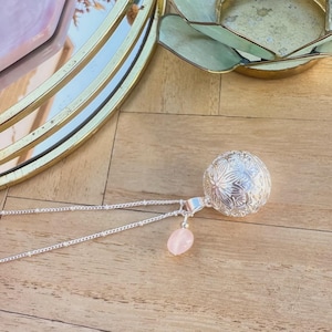 Peut inclure: Un collier en argent avec un pendentif sphérique finement travaillé et une petite pierre rose ovale. Le collier est posé sur une surface en bois, avec un plateau doré en arrière-plan. Le pendentif sphérique présente un motif floral.