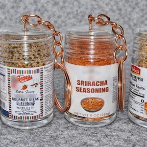 May include: Four clear spice jars with copper-coloured handles and chains. The jars contain various seasonings, including steak seasoning, Sriracha seasoning, and garlic pepper. Each jar has a label with the seasoning name and brand.