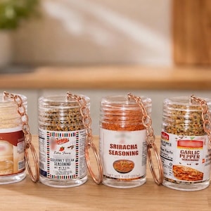 Könnte beinhalten: Vier klare zylindrische Behälter gefüllt mit verschiedenen Gewürzen, jeweils an einem kupferfarbenen Metallschlüsselanhänger befestigt. Die Etiketten zeigen Text, darunter Gourmet Steak Seasoning, Sriracha Seasoning und Garlic Pepper Seasoning.