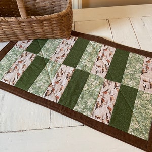 May include: A brown quilted table runner with a green and white patterned fabric. The fabric features a repeating pattern of birds and leaves.