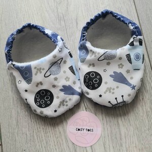 Può includere: Un paio di scarpine per neonati bianche con bordo blu e una stampa a tema spaziale. La stampa presenta razzi, pianeti, stelle e lune in nero e grigio. Le scarpine sono etichettate "Cosy Toes".