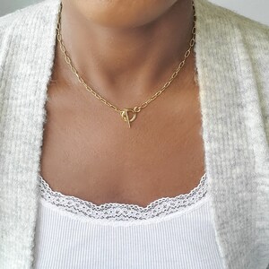 May include: A gold-colored necklace with an oval link chain and a T-bar clasp. The necklace is worn around the neck, and the clasp is centered. The background includes a white ribbed tank top and a light gray cardigan.