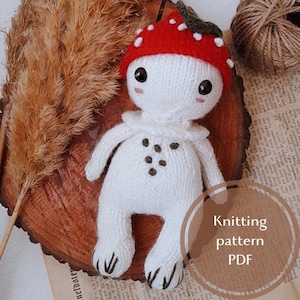 May include: A handmade white knitted mushroom doll with a red cap dotted with white spots. The doll has a ruffled collar and small brown spots on its chest. The doll is sitting on a wooden surface. The image also includes the text "Knitting pattern PDF".