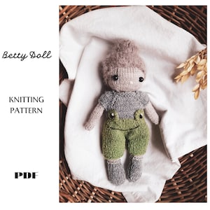 Può includere: Schema di lavoro a maglia per una bambola di lana chiamata Betty. La bambola indossa una maglietta grigia e pantaloni verdi con un disegno di rana. La bambola è sdraiata su un panno bianco in un cesto di vimini.