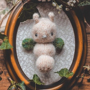 May include: A knitted plush toy in the shape of a butterfly with green wings, brown eyes, and a pink body. The toy is displayed in a wooden oval frame with a white floral pattern.