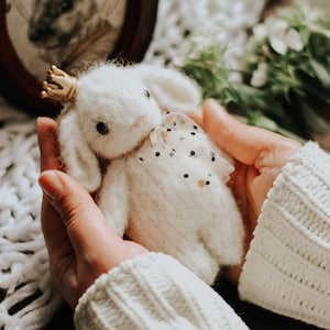 Puede incluir: Una muñeca de conejo blanca tejida a mano con una corona dorada y un lazo de lunares. La muñeca está sostenida en las manos de alguien, con un retrato de conejo enmarcado en el fondo. Un artículo encantador y caprichoso.