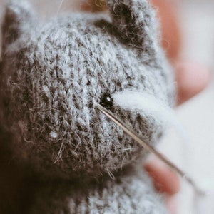 May include: A close-up of a gray knitted stuffed animal with a needle and thread being used to attach a white felt eye.