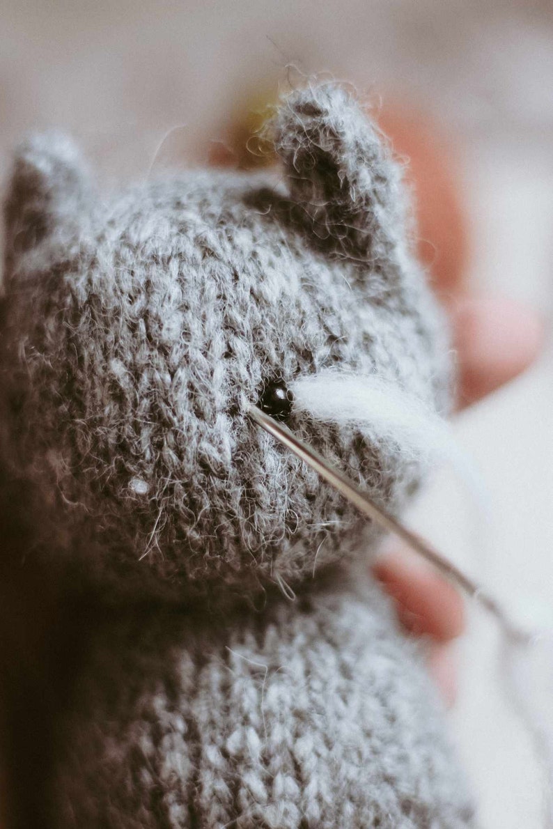May include: A close-up of a gray knitted stuffed animal with a needle and thread being used to attach a white felt eye.