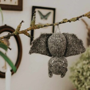 May include: A gray knitted bat hanging from a branch with green moss. The bat has a small, round body and large, pointed wings.