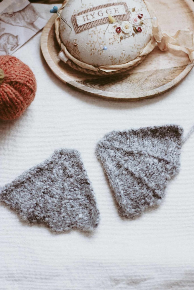 May include: Two gray knitted wings with a scalloped edge, lying on a white fabric surface. The wings are made of a soft, fluffy yarn.