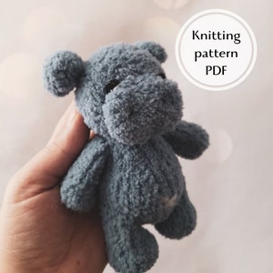 May include: A gray knitted plush toy hippopotamus. The toy is being held in a person's hand. The text "Knitting pattern PDF" is visible in a circle in the upper right corner of the image.