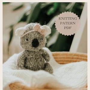 Puede incluir: Patrón de tejido de punto PDF para un koala de punto gris con una diadema rosa con lazo. El koala está sentado en una cesta de mimbre forrada con una manta blanca.