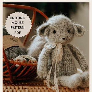 Puede incluir: Un juguete de ratón gris tejido con una cinta de encaje blanca. El juguete está sentado en una cesta de mimbre con ovillos de lana. La imagen incluye el texto "Knitting Mouse Pattern PDF".