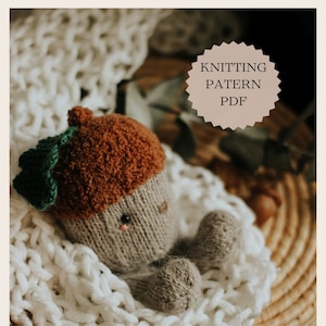Puede incluir: Un juguete de peluche con forma de bellota tejido, con una gorra marrón y detalles de hojas verdes, descansando sobre una manta de punto blanca. El texto "KNITTING PATERN PDF" se muestra en una etiqueta circular.