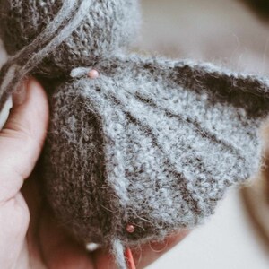 May include: A gray knitted stuffed animal with a long tail and wings. The animal is being sewn together with red pins.